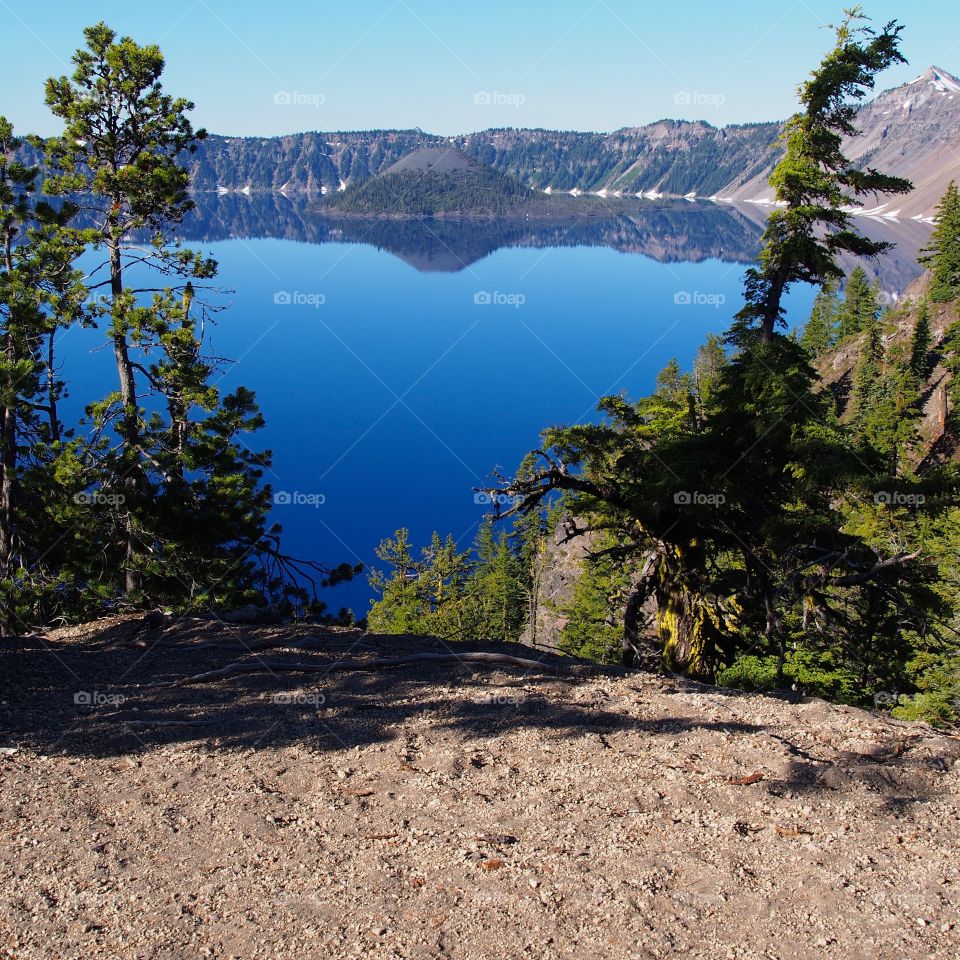 The rugged tree covered slopes of the rim of Crater Lake with Wizard Island across the lake in Southern Oregon on a sunny and clear summer morning.