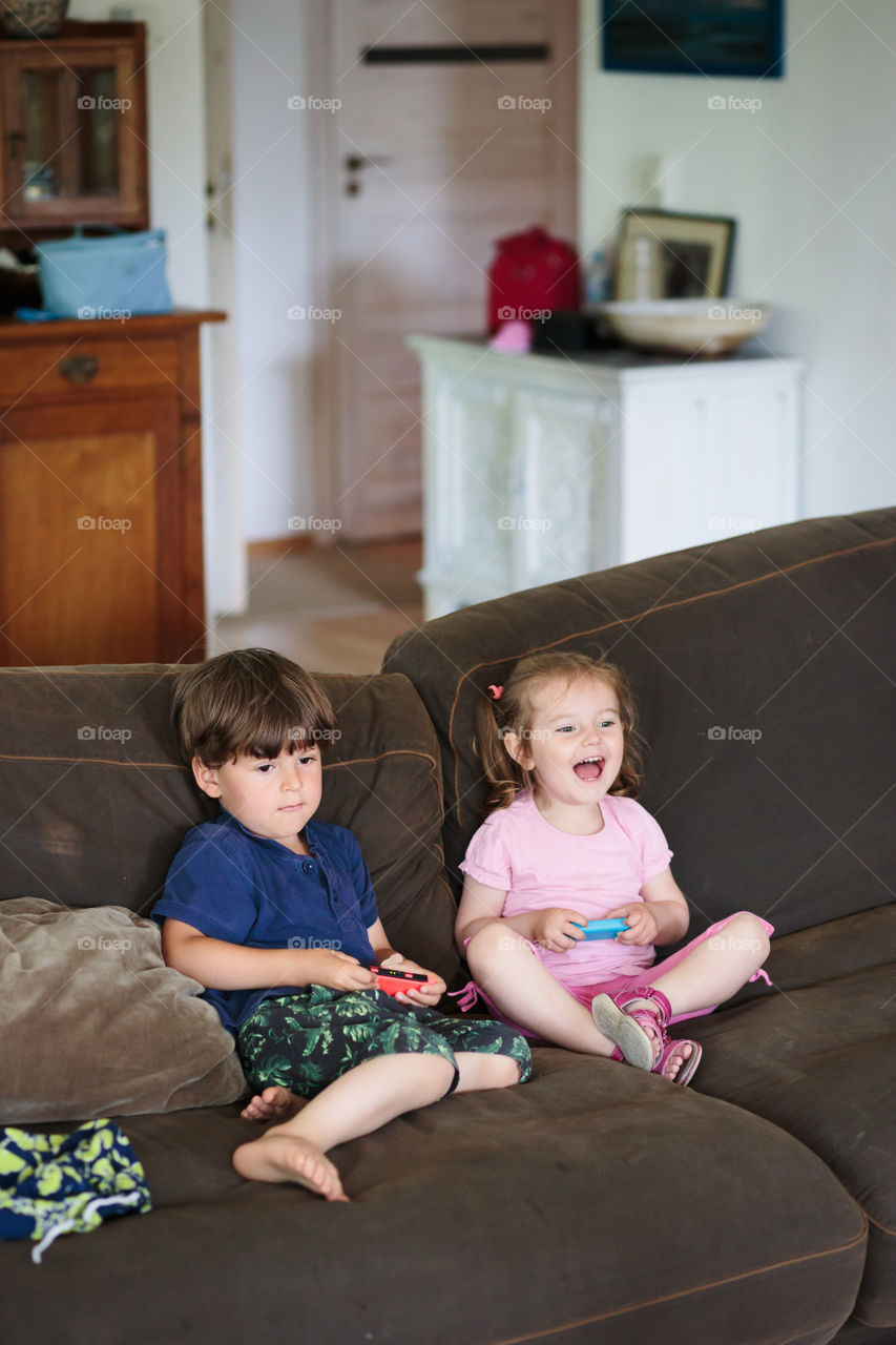 Happy smiling toddlers boy and girl playing video game sitting on sofa at home