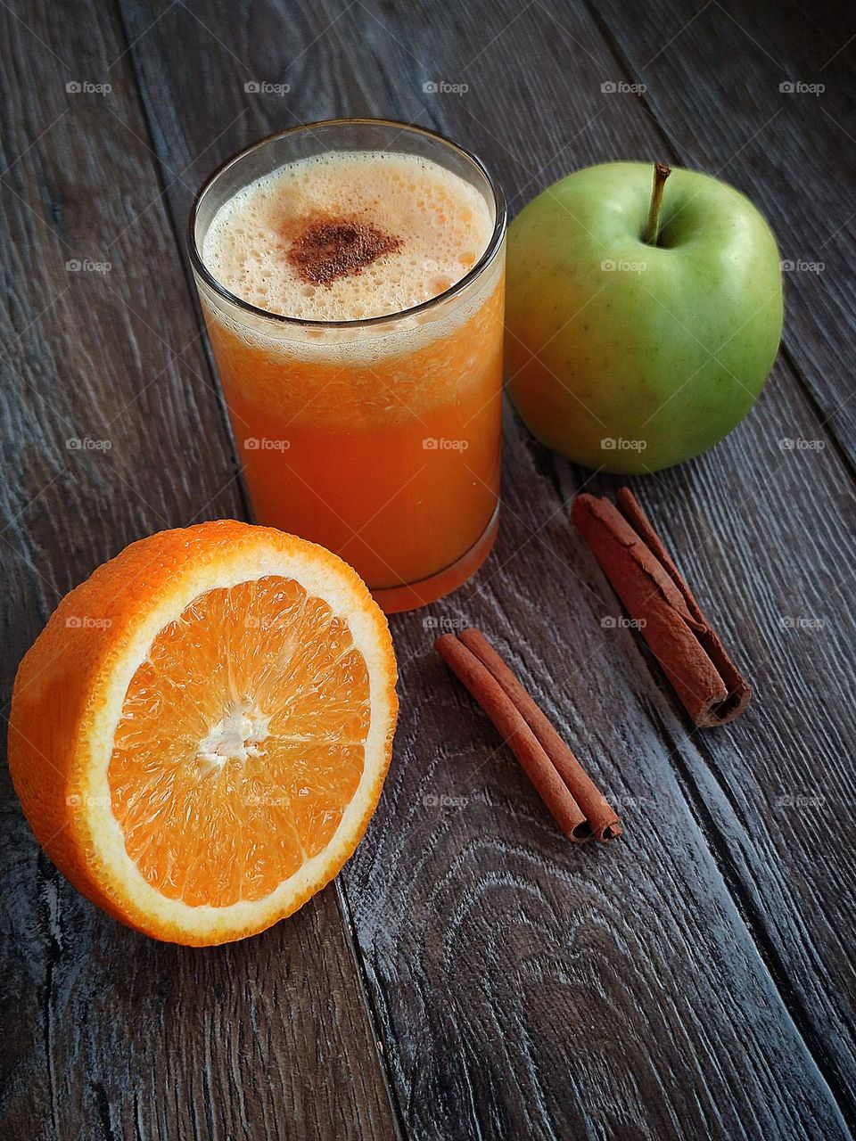 On a wooden surface stands a glass with an orange smoothie, the surface of which is decorated with ground cinnamon in the form of a heart.  Ingredients lie nearby: half an orange, a green apple and two cinnamon sticks