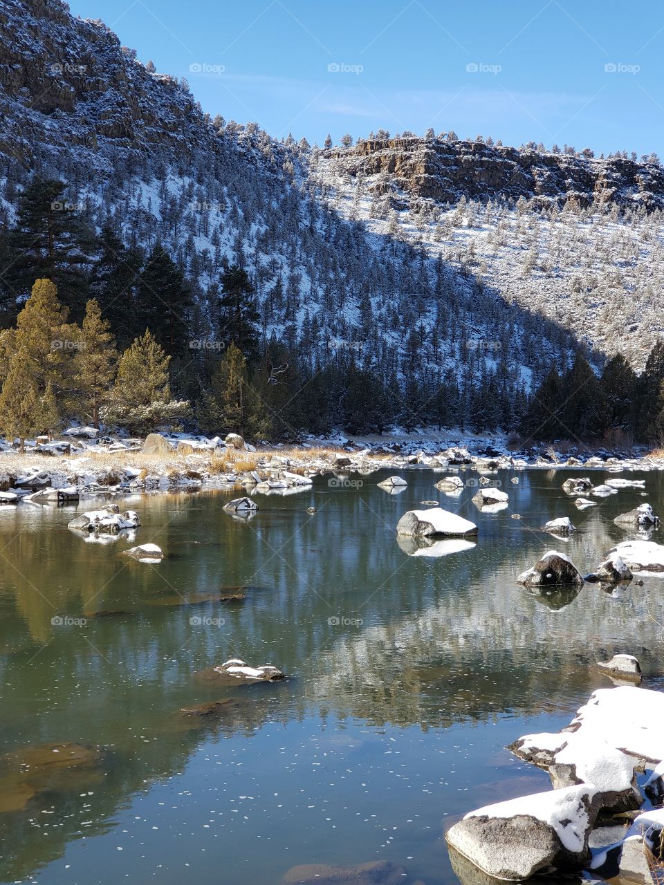 The Crooked River in Central Oregon flows through boulders and a canyon with a fresh covering of snow on a sunny winter day with blue skies.