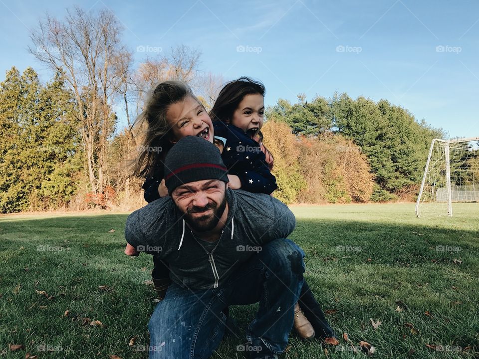 Dad with two cute little girls on his back playing, being silly, and having fun on the grass in the park. 