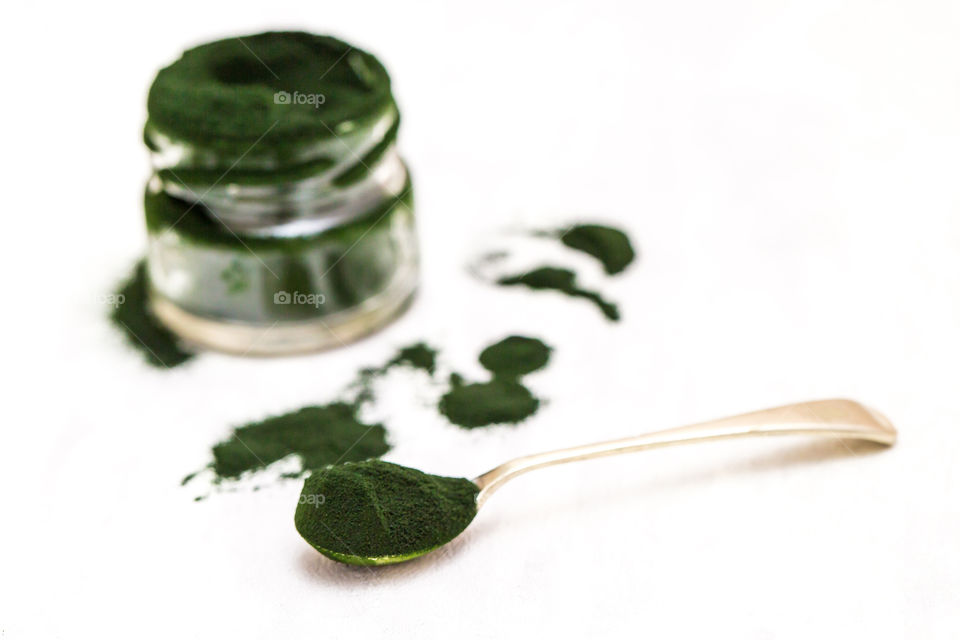 Product healthy green spirulina powder close up in glass jar with spoon on pure white background