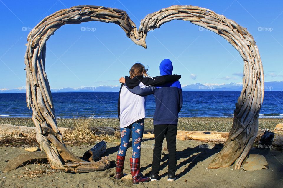 Two friends who have been friends since they were little are enjoying the view from their island home. From under the driftwood heart they can view the inky blue ocean, the majestic coastal mountains & the endless expanse of light blue cloudless sky!
