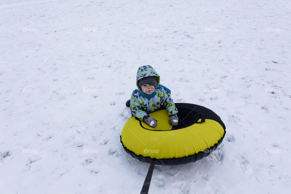 A small, carefree boy walks in the white snow in winter and rides a tubing in the park, near trees in the snow.