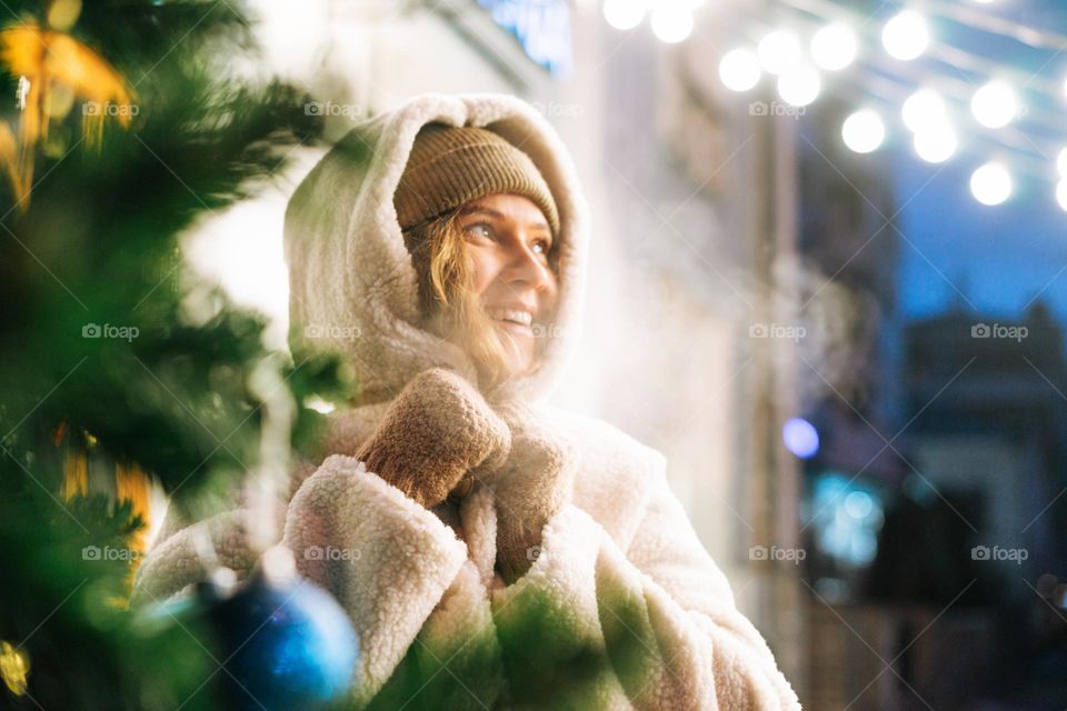 Young happy woman with curly hair in fur jacket having fun in winter street decorated with lights, Christmas fair 