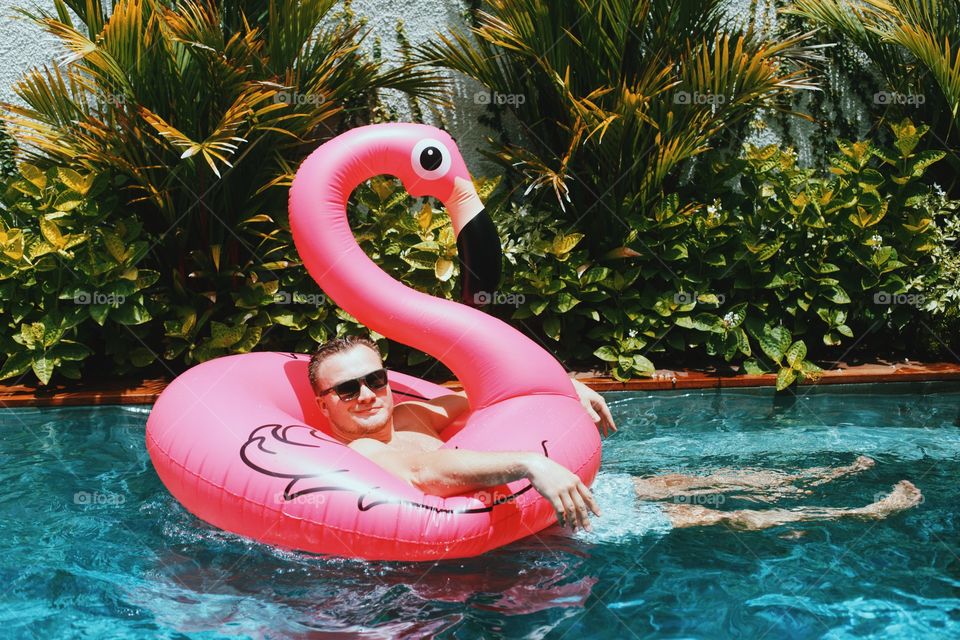 Guy in a pool on a pink flamingo floaty 
