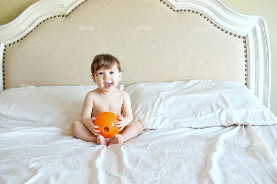 A smiling young toddler holding an orange ball while sitting on a bed with fresh clean white sheets 