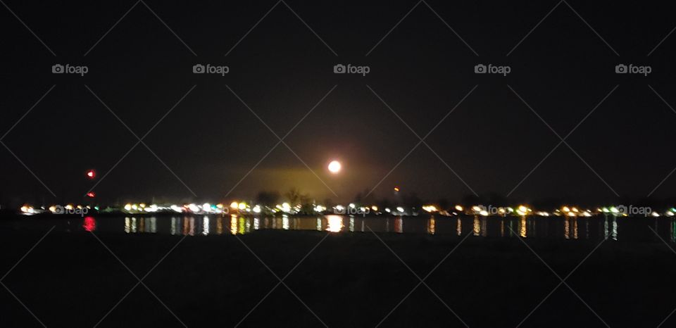 The town lights from a distance reflecting g over the lake with the shining moon in the center of the black sky