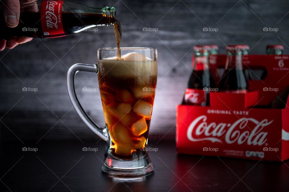 Backlit closeup of a glass bottle of Coca Cola being poured into a drinking glass 