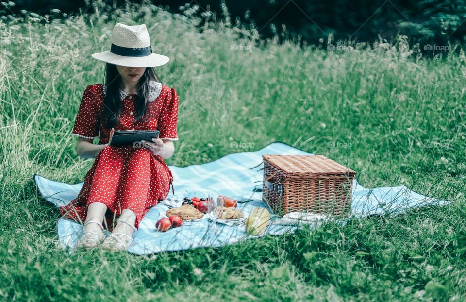 One young caucasian girl in a red dress and a straw hat sits on bedspreads with a wicker basket and fruits, browsing social networks in a tablet, relaxing in a park on a summer day, close-up side view. Picnic time concept.