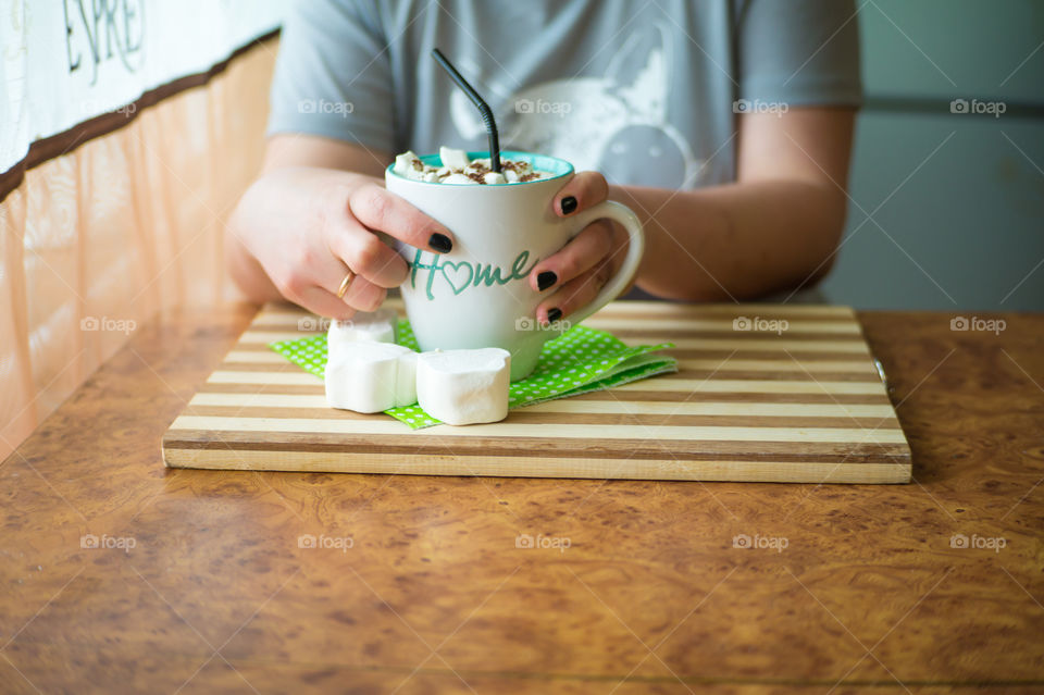 Black coffee with white melting marshmallows, sprinkled with chocolate chips with a black straw in a white mug with the inscription house in female hands.