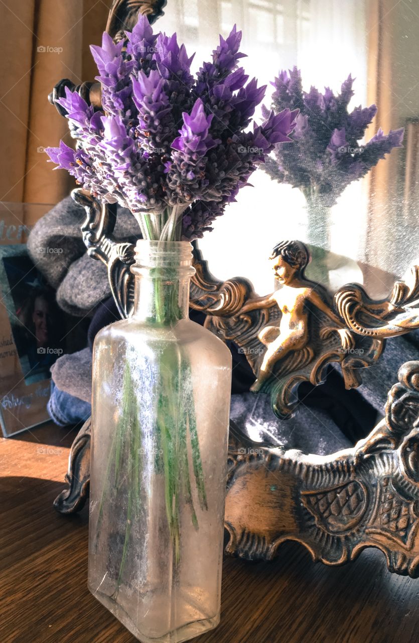 Bouquet posey of lavender flowers in an antique medicine bottle vase and sitting atop an antique wood dresser in front of an antique mirror. Bouquet reflected in mirror