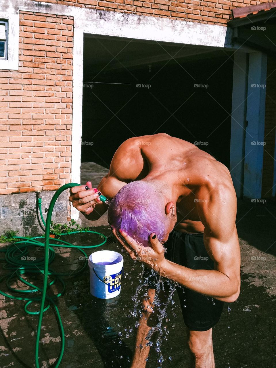 Man with purple hair washing his hair with a water hose. Painted black nails and colorful hair.