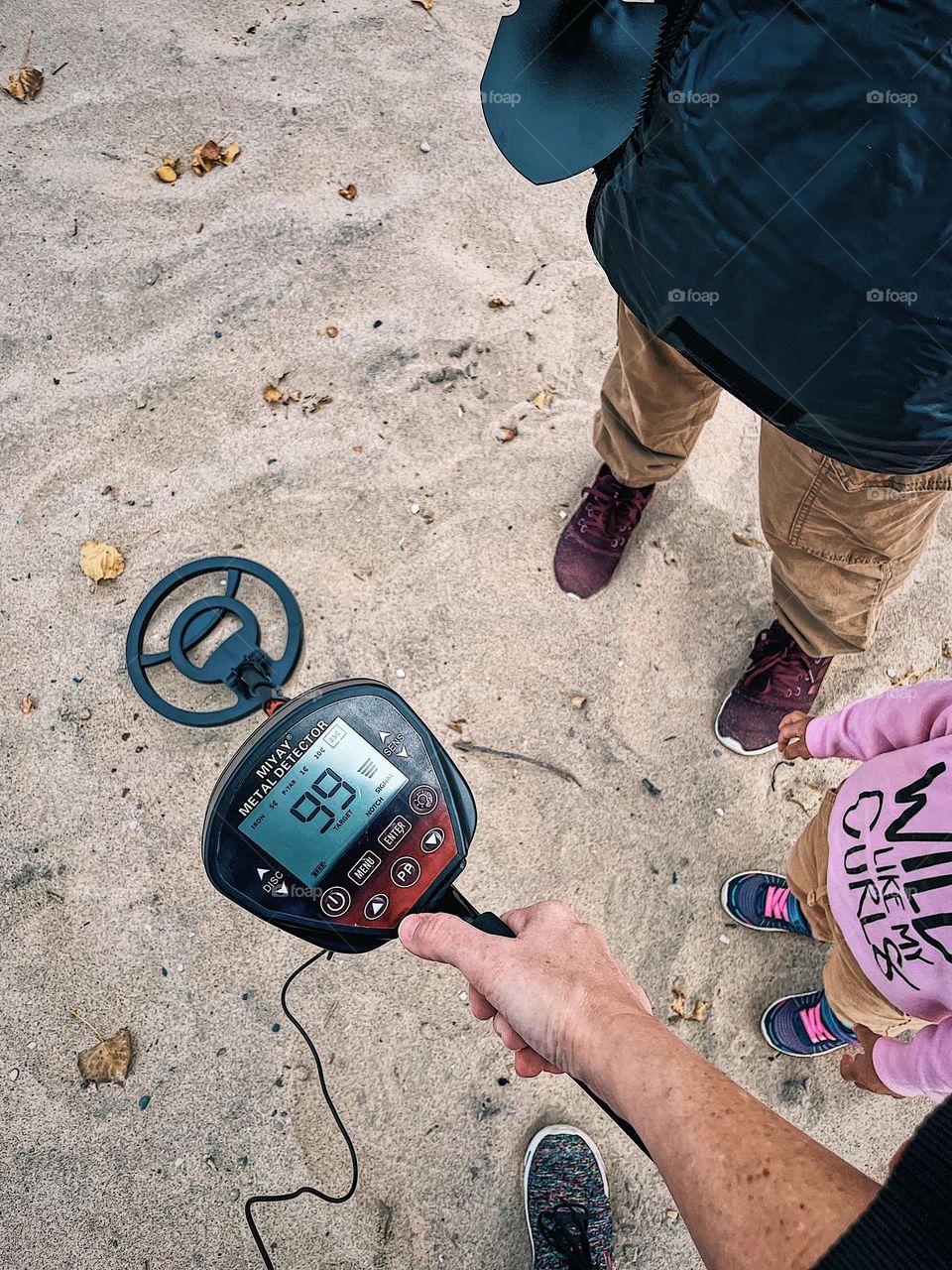 Family looks for treasure on the beach, finding treasure by the shoreline, family beach activities, spending time with family doing unusual activities, woman’s hand holding metal detector, searching the beach for treasure with family