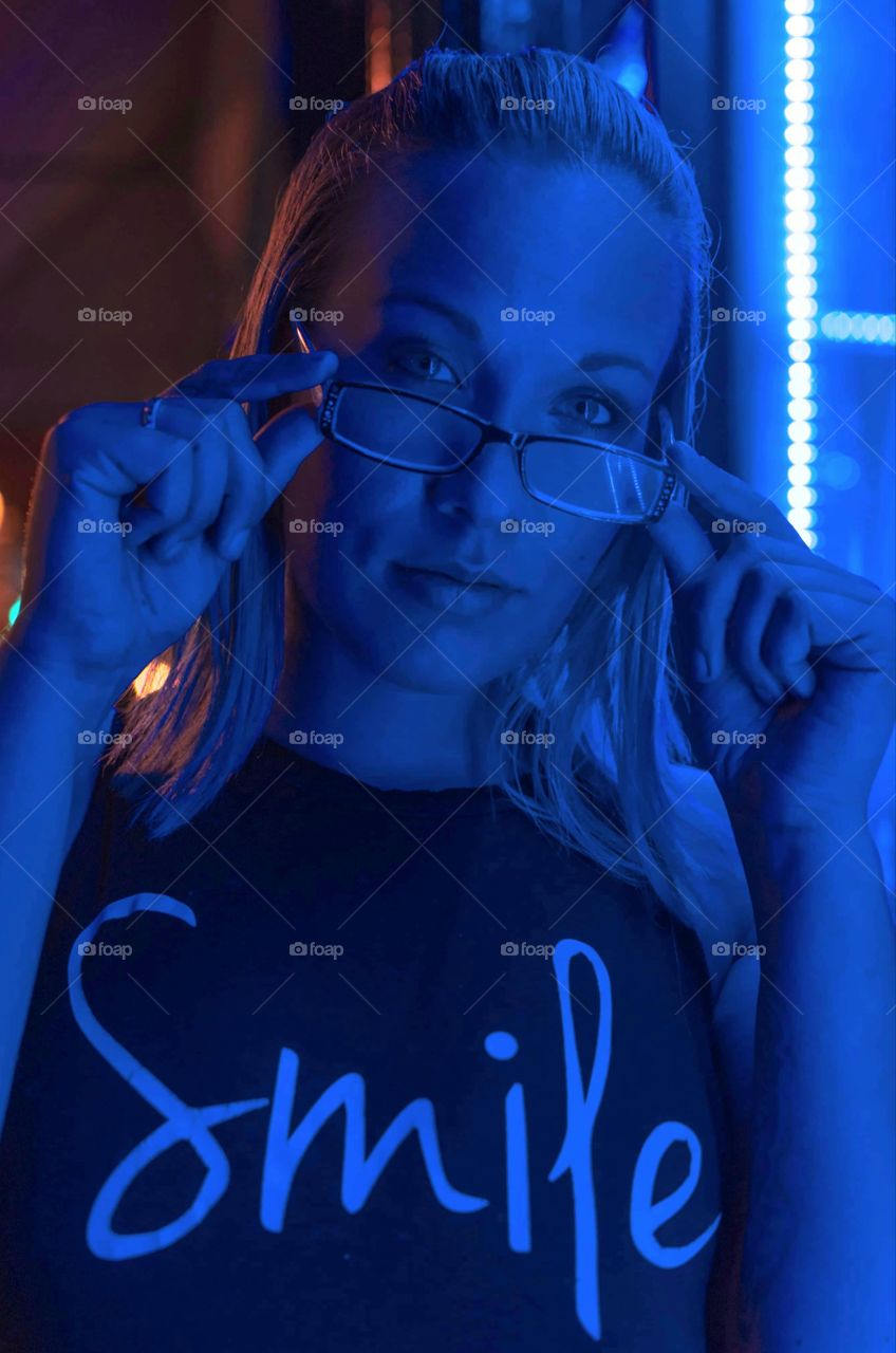 night portrait image of a beautiful girl wearing a t-shirt that says smile pulling her glasses down on her face while looking into the camera all in blue light from store windoe