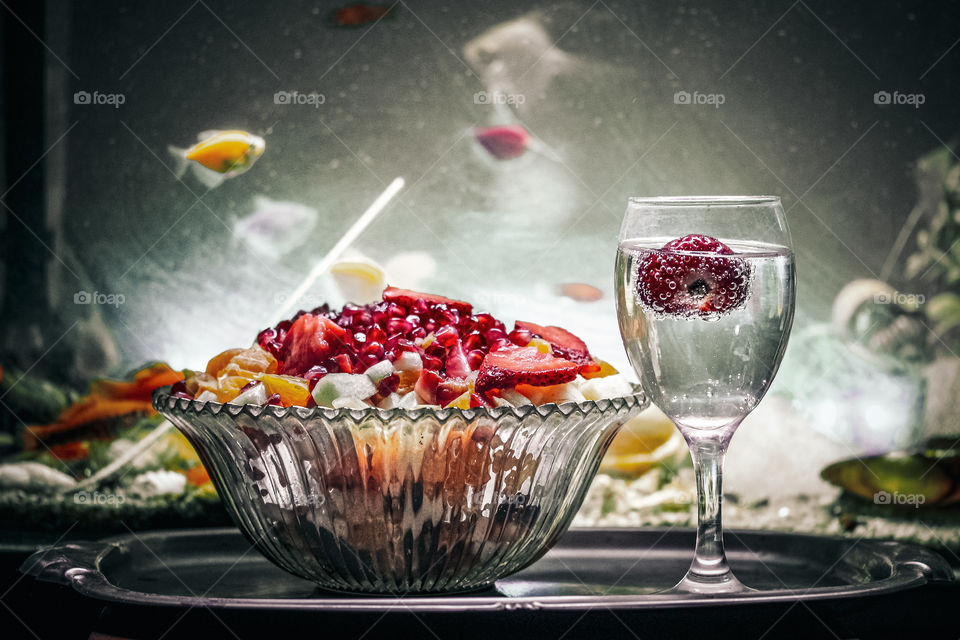 A fruit salad and a glass of mineral water with strawberry at the water and aquarium fish swimming in the background in the aquarium