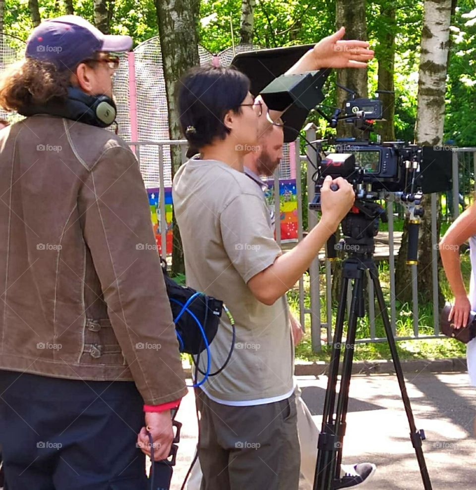 Summer in the city. In the park, among the green trees, a movie is being shot. In the main plan is a cameraman waving to the actors.
