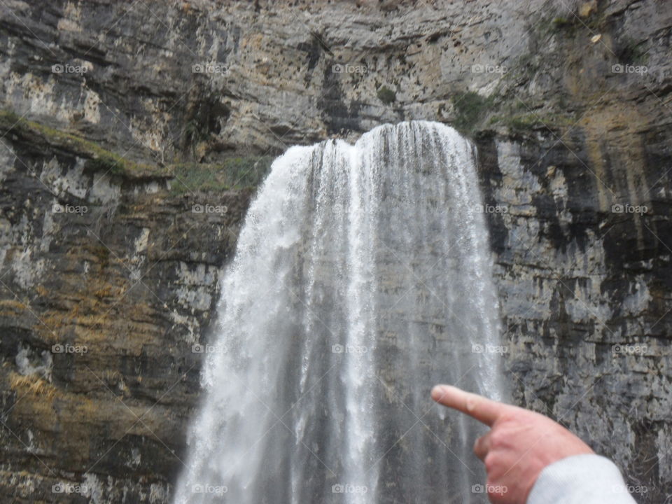 It is a waterfall in the middle of the mountains. The water practically comes out of the rocks. My partner's hand also comes out, pointing a finger towards the waterfall.