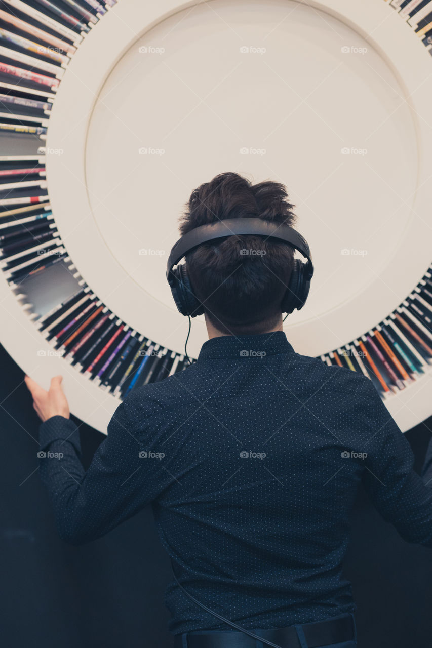 Young man listening to music through headphones standing next to bookshelf with recordings
