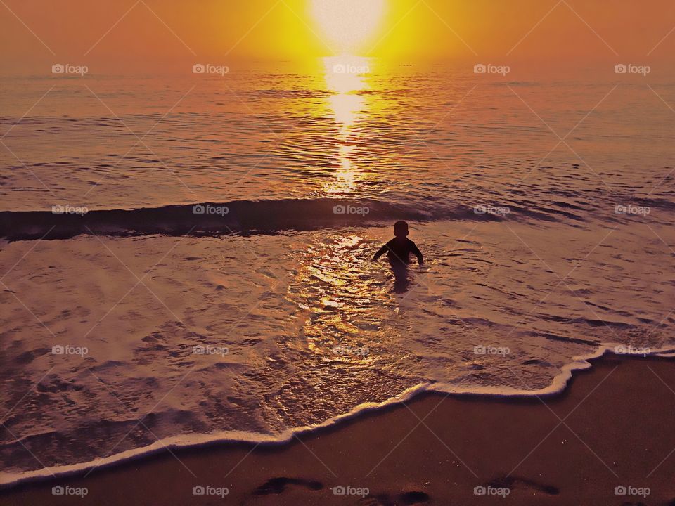 A small boy playing in the ocean the summer sunset bathing him in golden light.