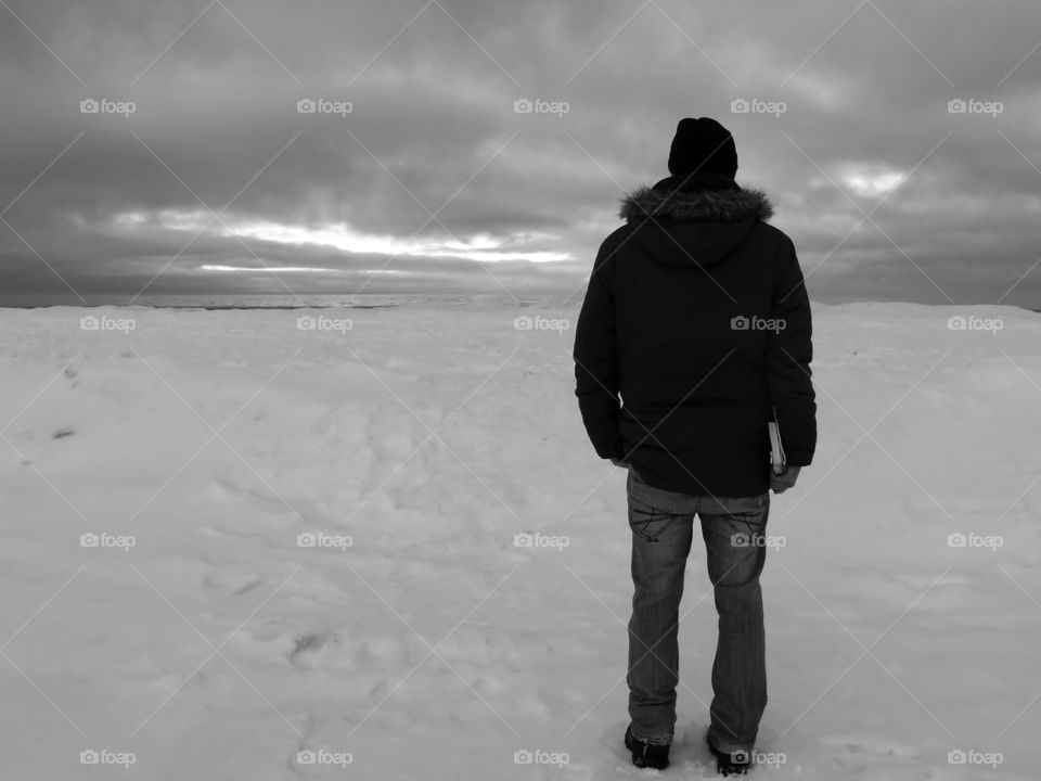 Black and white rear view shot of young man in warm clothing standing on snow covered beach in Jūrmala, Latvia.