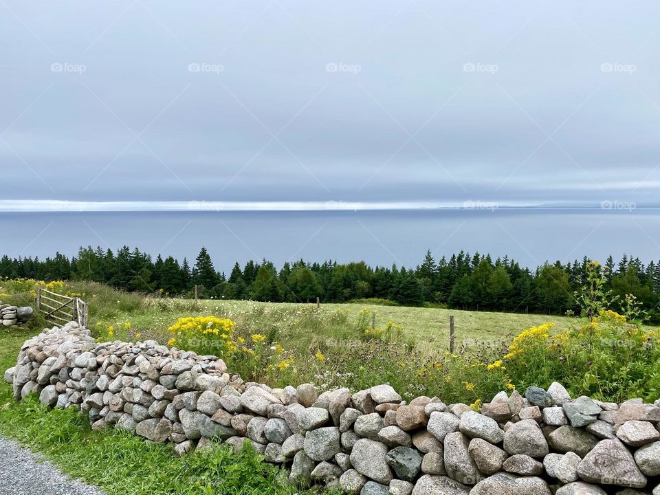 A stacked stone fence surrounding a field atop a hill overlooking the water on a grey overcast day