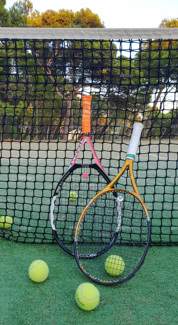 Attributes of the game of tennis.  Two tennis rackets and tennis balls on an outdoor court in summer near the net against the backdrop of a pine forest and the sea