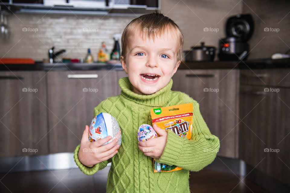 A boy in green sweater holds sweets and smiles. Happy childhood. Kid loves kinder and m&m. Favorite food