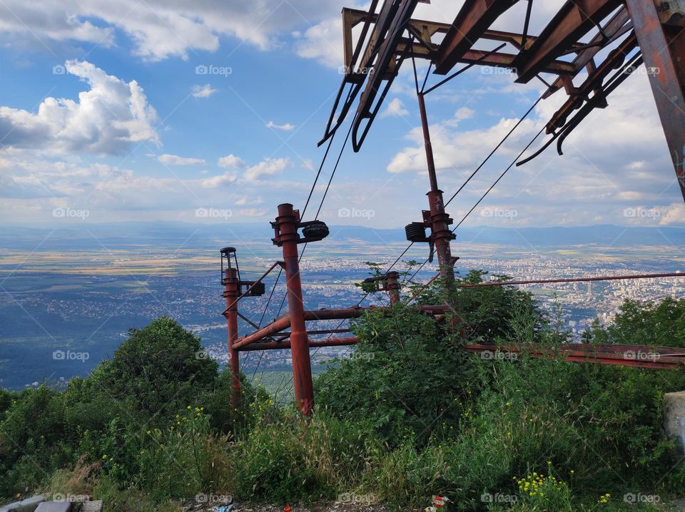 An old abandoned metal construction for a ski-lift in the mountain with a view of the capital city of Bulgaria, with beautiful skies full of clouds and plants around the metal