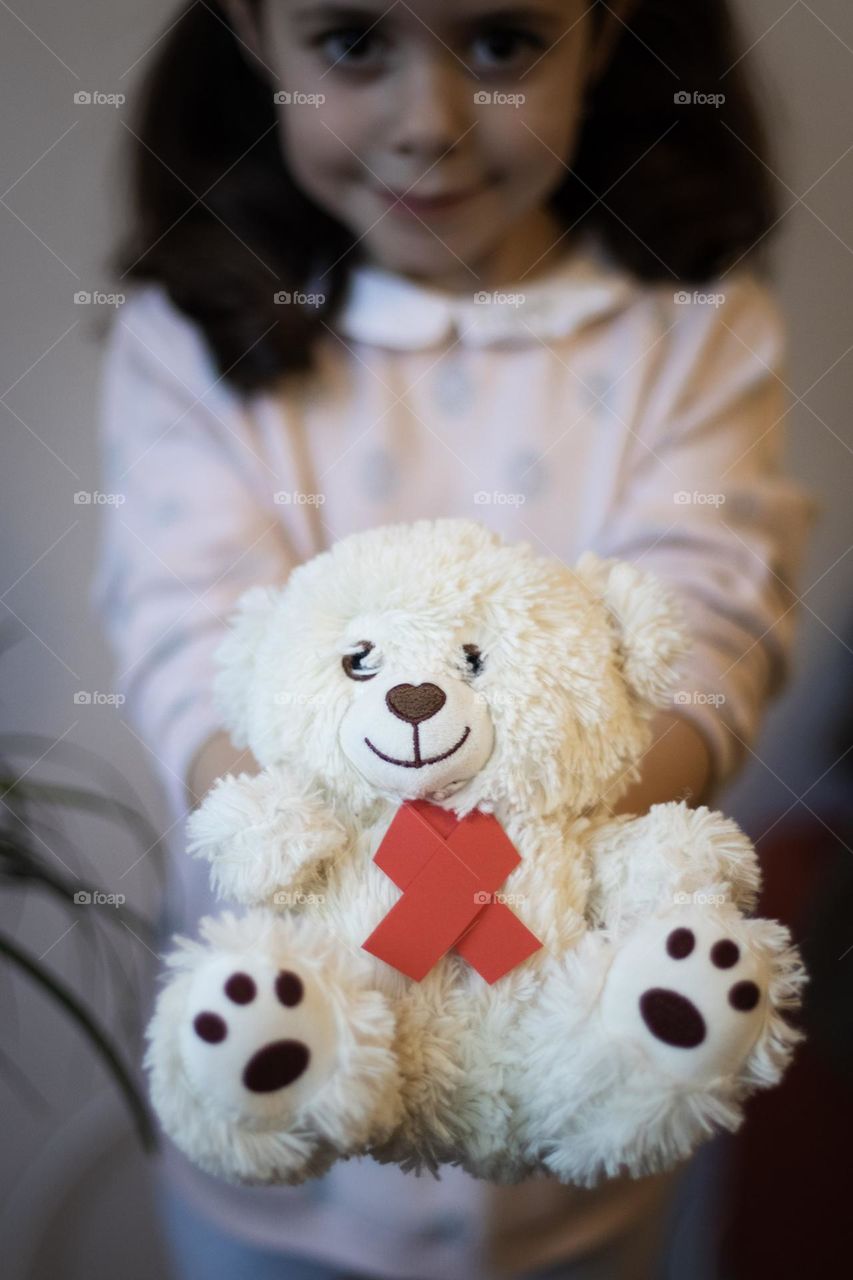 One beautiful caucasian brunette girl is holding a white fluffy teddy bear with a red paper ribbon tie on her chest, stretching it forward, vtd side closeup. Children's HIV and AIDS day concept.