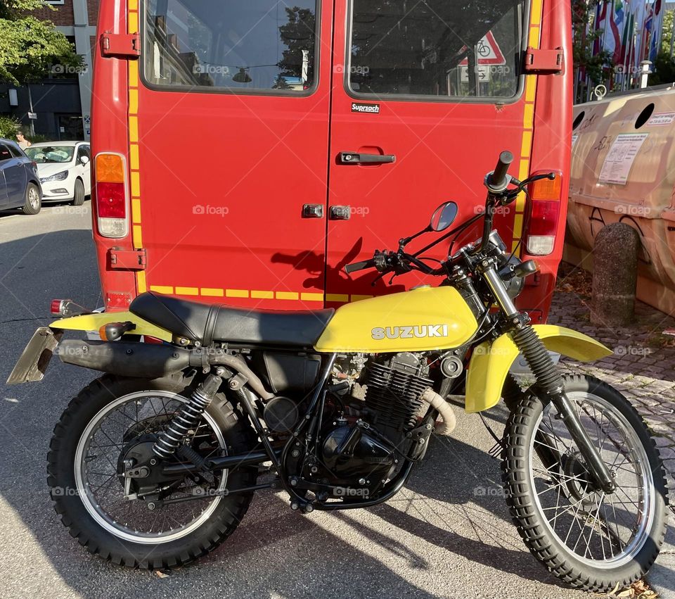 A vibrant yellow Suzuki motorcycle is parked behind a striking red van on a street in Munich, Germany. The motorcycle stands out, contrasting against the red van's deep hue. The scene captures a mix of urban energy and bright colors.