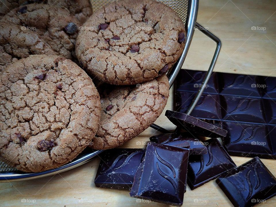 In a metal sieve are chocolate chip cookies with chocolate chips.  Nearby are black pieces of chocolate and half a chocolate bar.