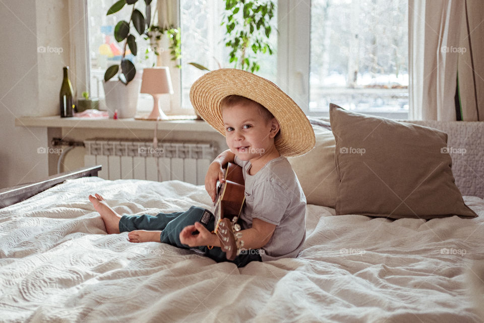 Little boy playing ukulele guitar in straw hat at home