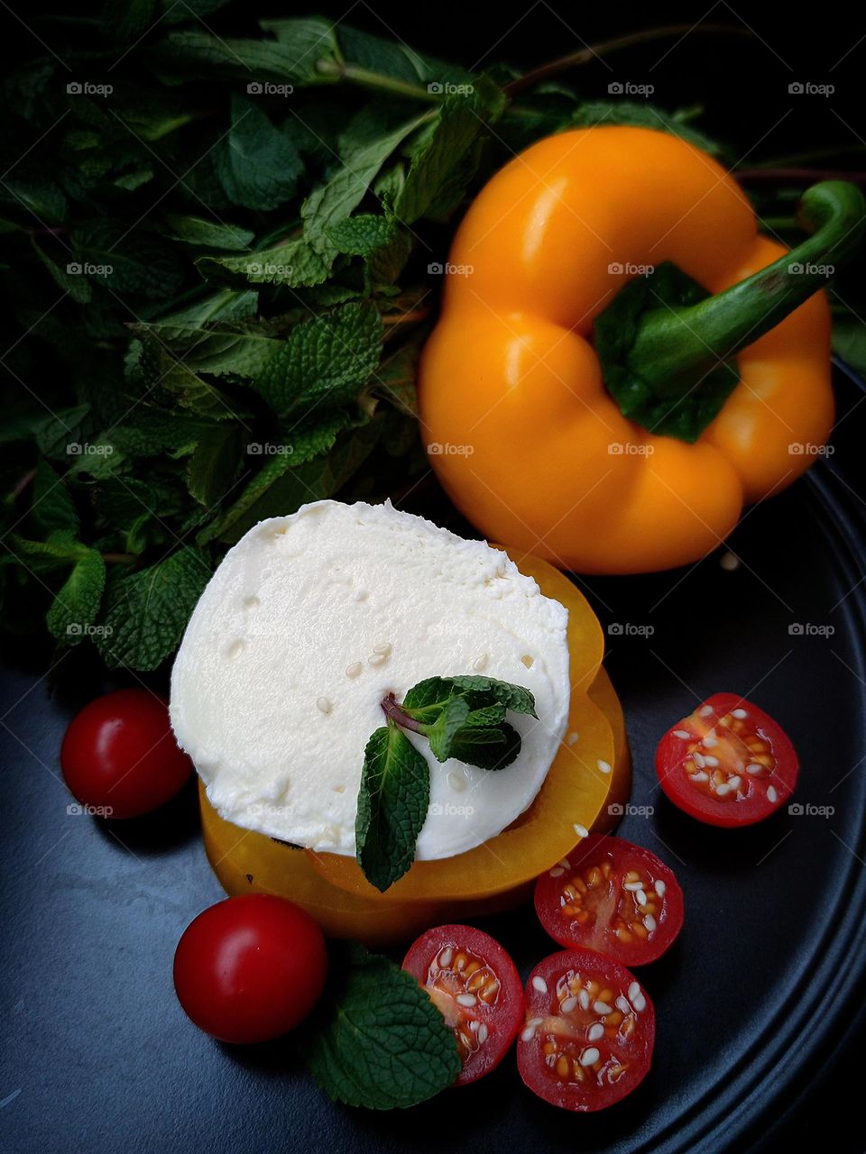Still life: white burrato cheese lies on mugs of yellow bell pepper. Nearby are cut red tomatoes and red tomatoes, yellow bell pepper, which closes a bunch of green mint. Cheese and tomatoes sprinkled with sesame seeds. Black background