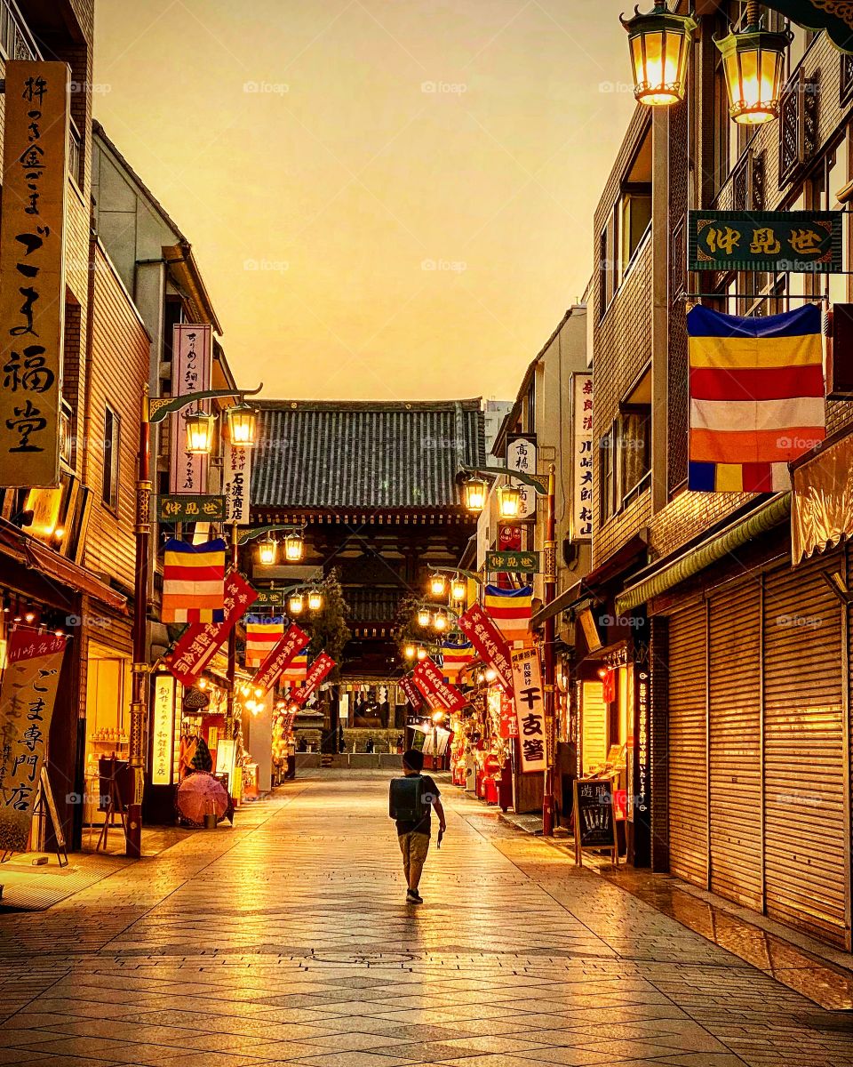 A child walks home after school at sunset, street lamps switched on and shops leading up to a temple closed or closing for the day. 