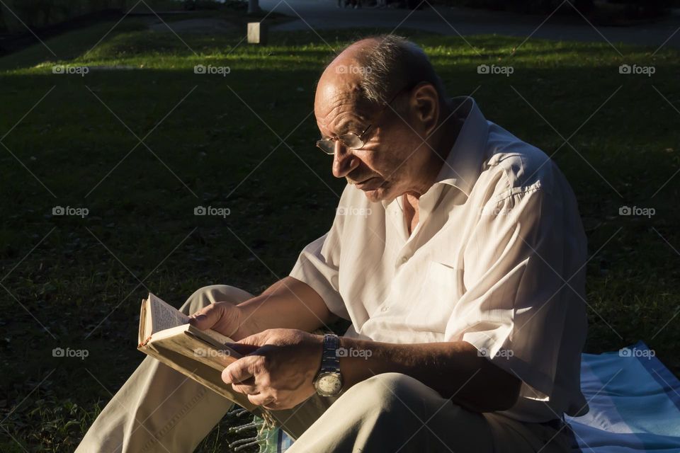 An elderly man of retirement age with wrinkles sits on the grass in the park and reads a book.