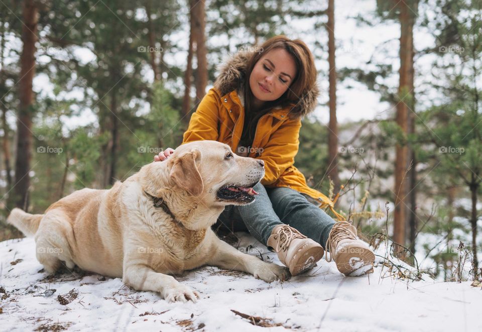 Young smiling woman in yellow jacket with big kind white dog Labrador walking in the winter forest