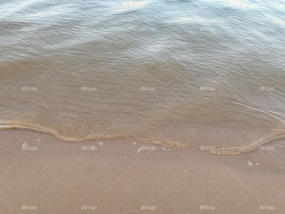 This image captures a close-up view of gentle waves lapping against a sandy beach. The water appears calm, and the shoreline is smooth with small bubbles forming at the edge where the water meets the sand, a peaceful and serene beach scene.