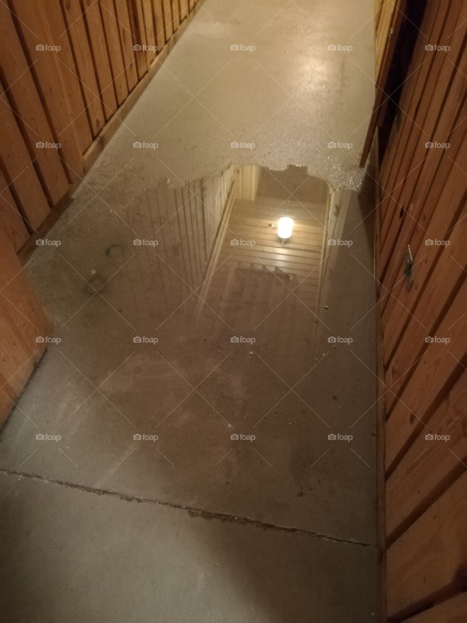 The air conditioner is broken and let the water fall down on the dirty floor of the cellar. The wet surface makes reflections of the ceiling, the electronic device and the lamp. One door is slightly ajar.