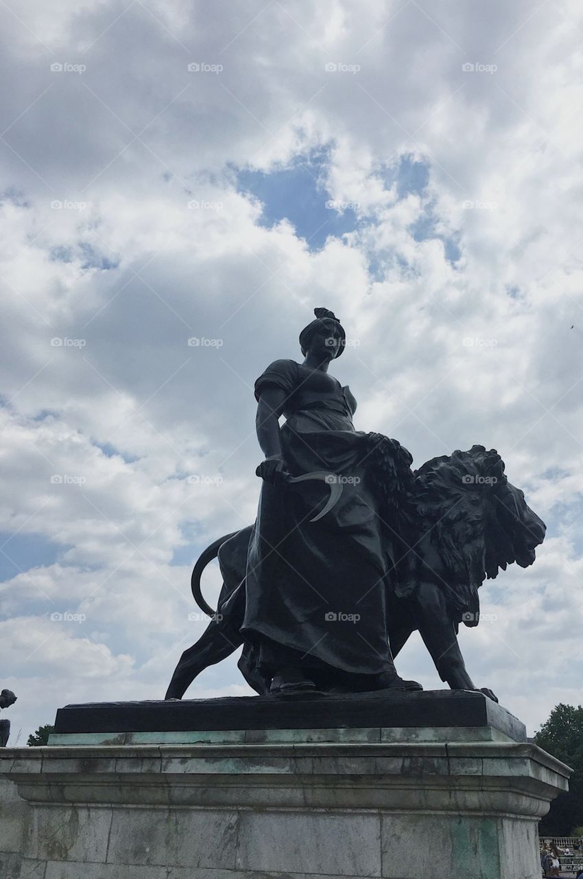 A black statue outside of Buckingham palace, a cloudy sky is behind her, she stands with a lion.