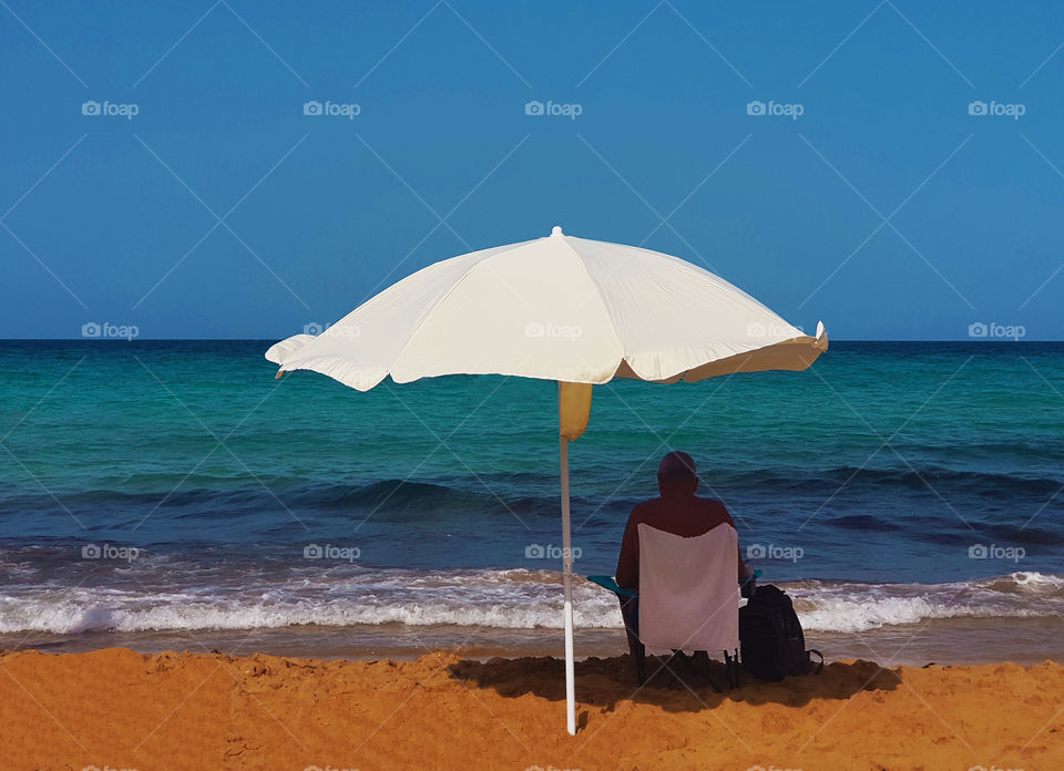 man on beach under white umbrella and blue sky surrounded by brilliant blue sea