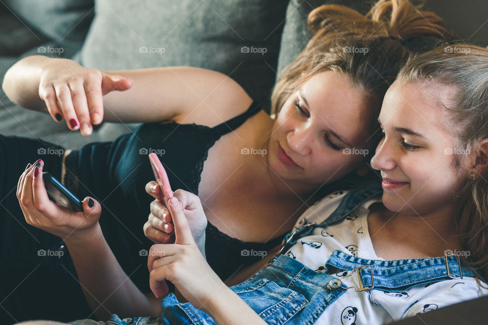 Young women using mobile phones watching music clip, texting, messaging. Teenagers using the smartphones, sitting on sofa at home. Using technology devices. Girls wearing summer clothing