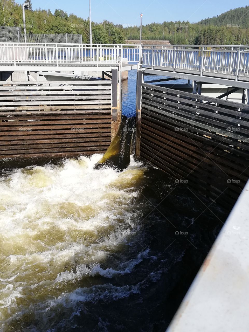 A waterway enclosed by gates, used for raising and lowering boats between levels, is open to let the water rush bubbling strongly. A metallic footbridge is empty and divides in two for a moment.