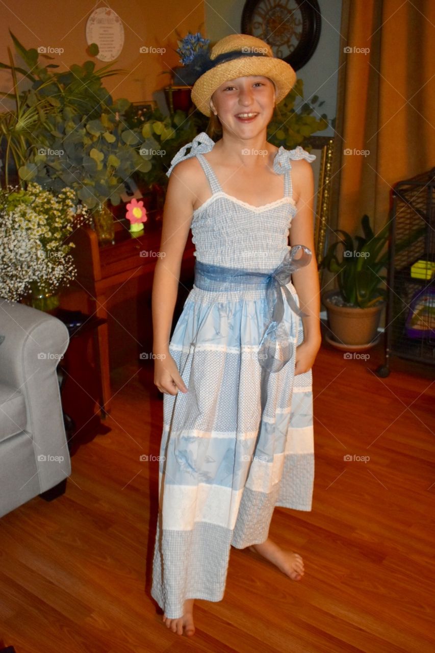 A bright and happy young girl stands barefoot in a hand made blue dress smiling at the camera. She is surrounded by eclectic furniture and decor, flowers and greens, on wood flooring. 
