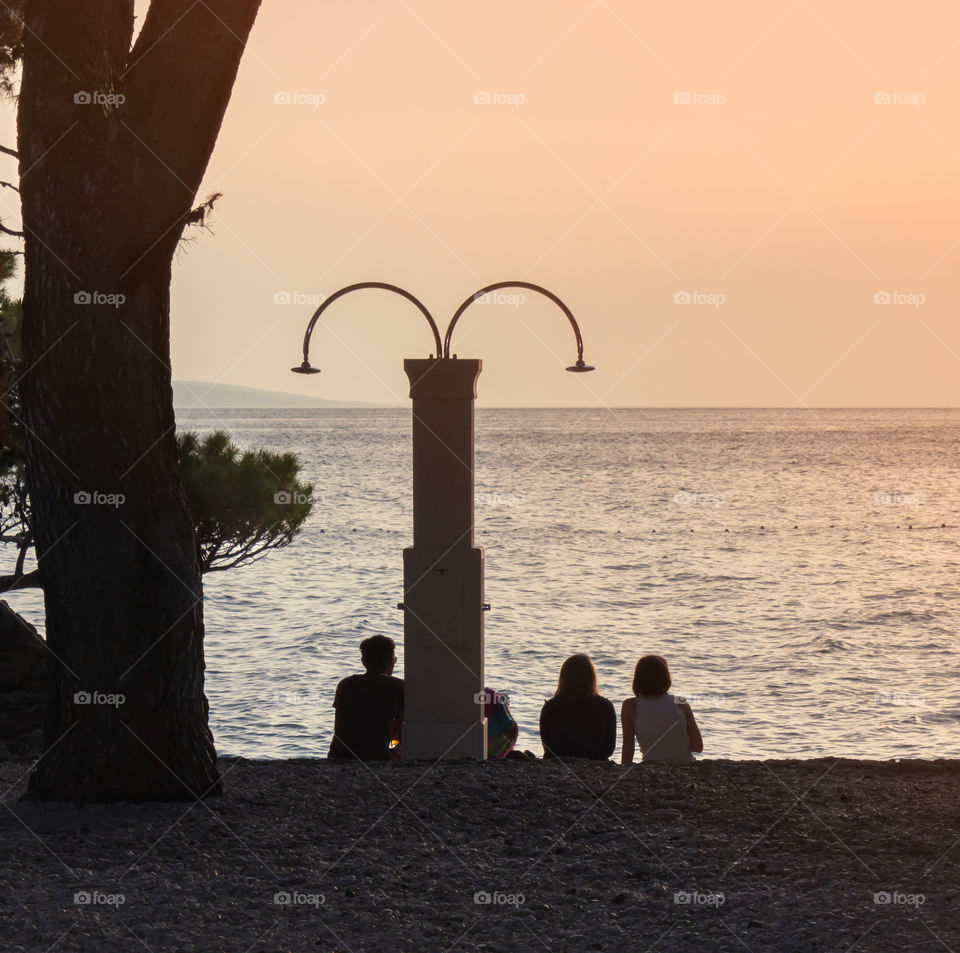 Silhouettes of young people sitting on a pebble beach in the evening, admiring the sunset. Croatia, Dalmatia, Brela, sunset in the sea on the beach of Punta Rata