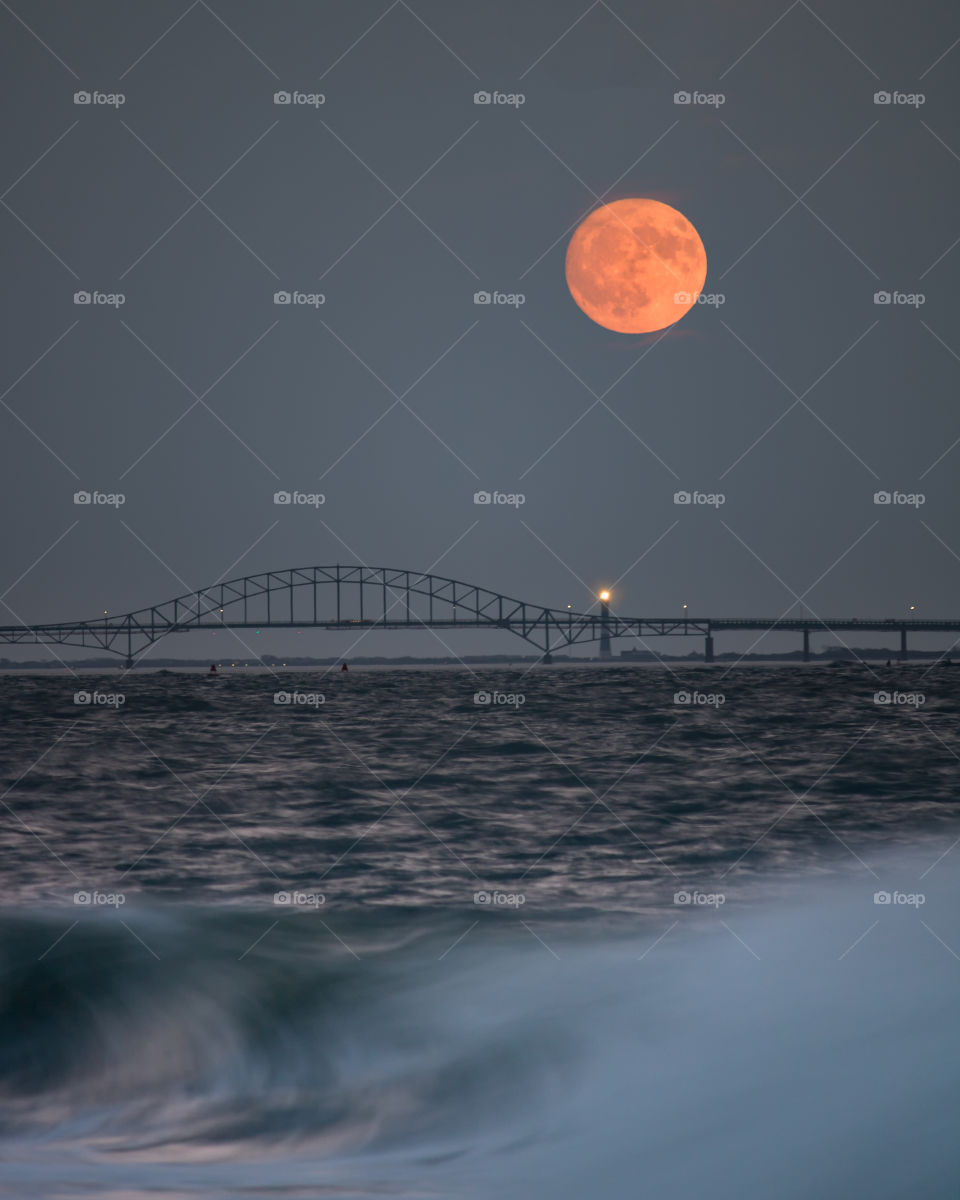 An orange bright full moon rising over a beautiful bridge and lighthouse in the distance. 
