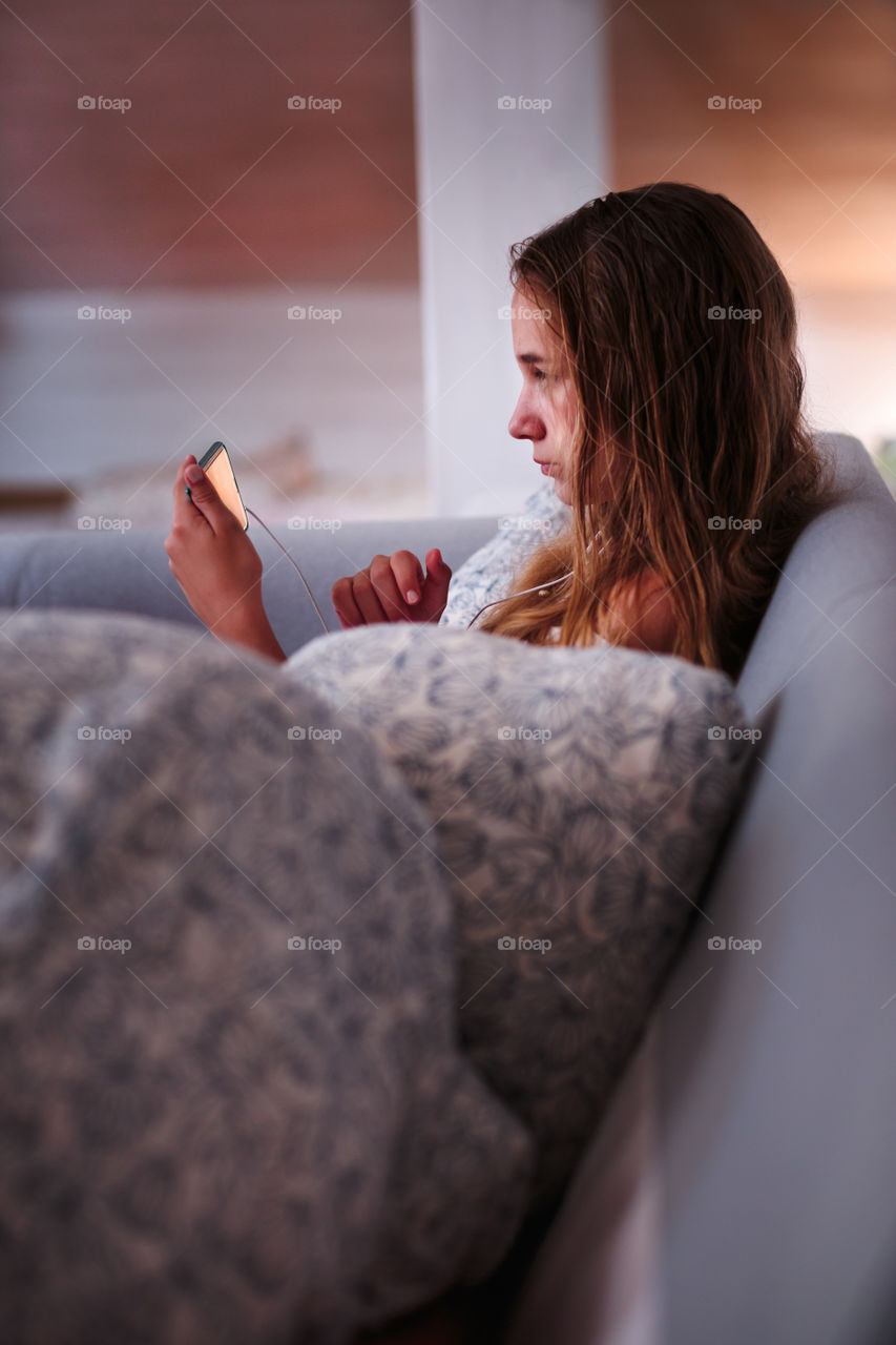 Teenage girl watching video online on smartphone sitting on sofa at home
