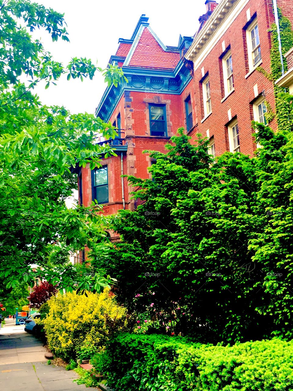 The unusual Fort Greene, Bklyn brownstone w multiple details: an adjacent tower w a steeple shaped rooftop, delicate stucco decor below the offset level of the tower in blue approx. complimentary to the red of the facade. 2019. Hypnotic Productions.
