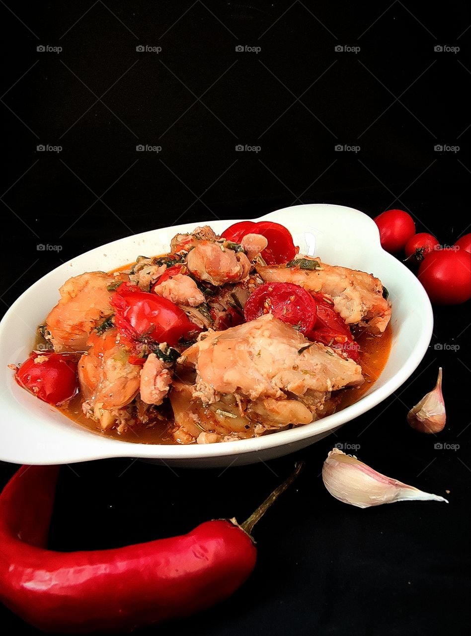 stewed meat with spices and cherry tomatoes in a white plate.  Nearby are two cloves of garlic, chili peppers and cherry tomatoes.  Black background