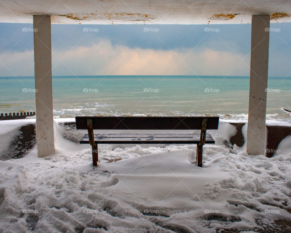 A view of a bench surrounded by snow looking out on to the sea and snow clouds in the sky - Bottle Alley, Hastings UK